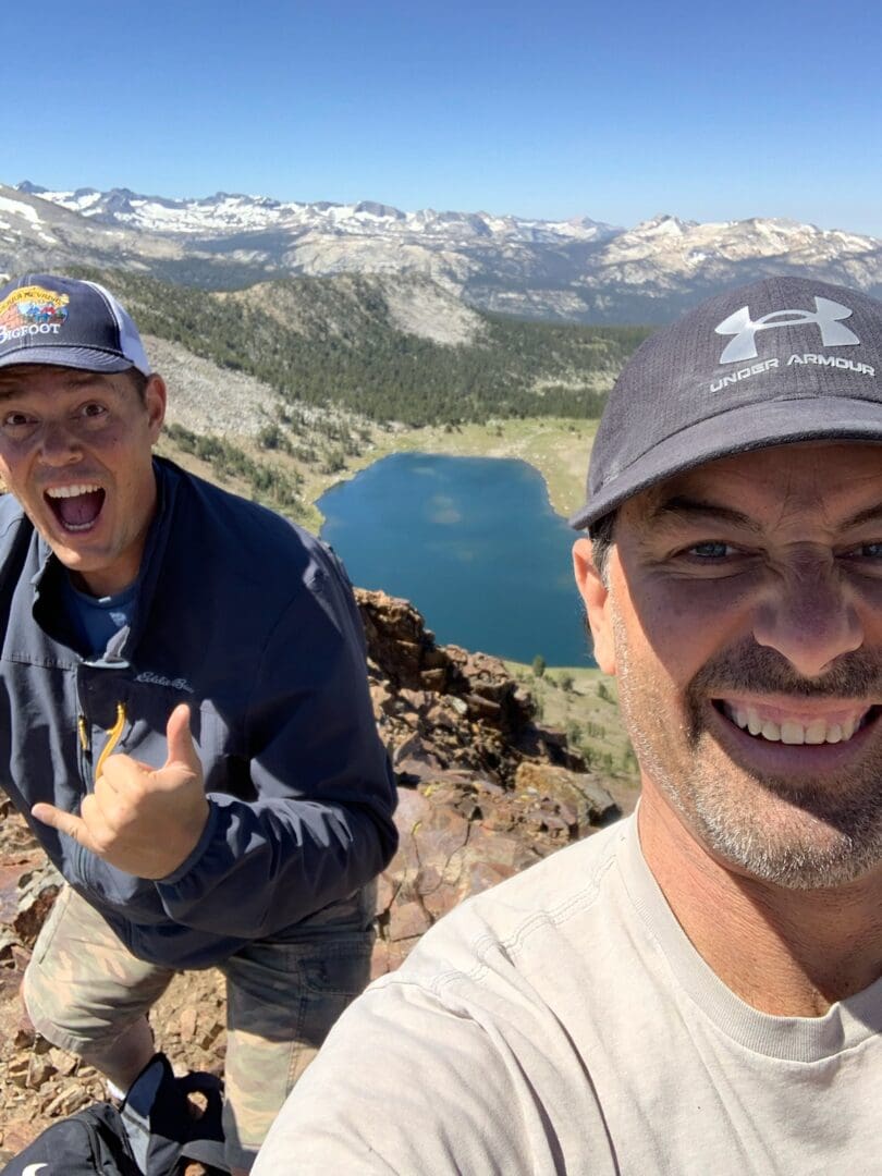 Two men smiling on mountaintop, lake view.